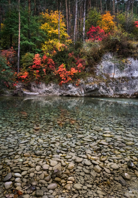 Color photo of a shallow clear river with a stony bottom and a forested steep riverbank on the other side with many trees in bright autumn colors. 
