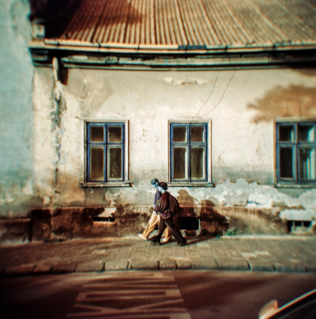 The photograph captures two people walking along an old, weathered wall with faded plaster and peeling paint. The building has a corrugated roof and several worn blue-framed windows that add a touch of symmetry to the scene. The soft focus and vignetting, characteristic of the Holga camera, create a dreamy and nostalgic atmosphere.

The colors are heavily shifted — the sky and walls glow with warm amber tones, while the shadows and windows take on surreal turquoise-blue hues. This unusual palette, produced by Lomochrome Turquoise film, transforms an ordinary street moment into something dreamlike and cinematic, as if reality itself were subtly misaligned