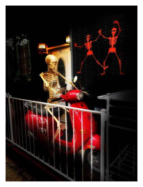 Photo of a life-size Halloween skeleton sitting astride a red Vespa motor scooter, which is parked inside a gated entryway in front of an apartment door.   Two illuminated lanterns hang above the lintel. To the right of the door, two flat orange skeletons, arms raised as if dancing, are affixed to the metal grid of a closed storefront gate. Filters have been applied so that the scene is darkened and the skeletons and Vespa glow softly against the shadows.