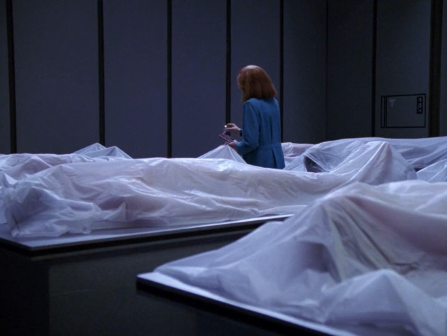 Star Trek The Next Generation scene. Pic 1: Crusher stands alone in a large storage room filled with many plastic bagged dead bodies laying on tables. Pic 2: Close up on Crusher looking serious, closed caption reads: [SHEET RUSTLES]
Pic 3: Camera shot further out, showing three of the bagged bodies now sitting straight up behind Beverly.  
Picture 4: View panned out even further to show the larger room and all bodies previously laying flat are now sitting straight up.  