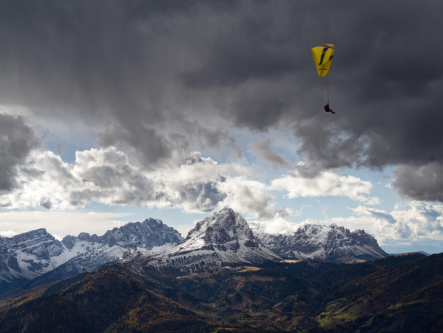 View as seen from the Kronplatz in South Tyrol. A paraglider with a yellow parachute is flying above the Dolomite mountains under a cloudy sky. Snow-capped peaks and green valleys are visible in the landscape.