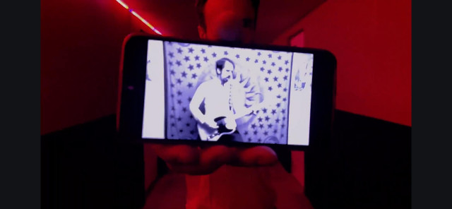 Schwarz-Weiß Darstellung eines Gitarristen mit Telecaster im weißen Hemd auf dem Bildschirm eines Handys, gehalten von einer Person in weißem Hemd, die in einem rot beleuchteten Gang steht.