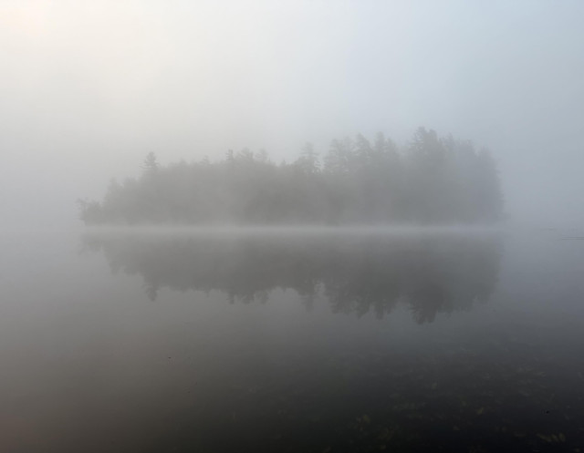 Ethereal photo of a small tree-covered island on a fogged-in section of lake with a white line of mist along the water’s edge; the bottom half of the shows the reflection of the island in water that darkens toward the bottom edge, faintly revealing a shallow, rock-covered river bottom; the top half is pale blue sky with a yellow-pink glow in the left corner from the invisible sun. 