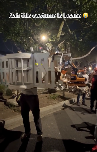 Night on a street in LA. A person wearing black pants and a white shirt has covered almost the entirety of their upper body in an incredibly elaborate replica of the White House with the destroyed East Wing. There is even a small model excavator as was seen in news photos.