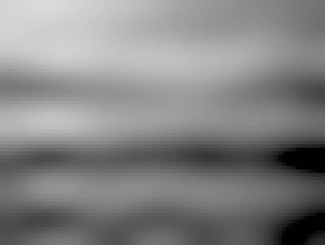 Large rocks and old cement slabs are seen along the shoreline of a lake. The smooth glass like water reflects the trees along the distant shoreline and soft clouds in this black and white image.