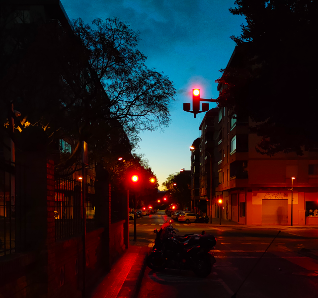 Photo of a street at dawn. Two symmetrical rows of short buildings fugue towards the center of the picture, leaving the sky as a centered pale blue triangle, with a bit of yellow on its lower vertex. The city is still in shadow, lit by red traffic lights.