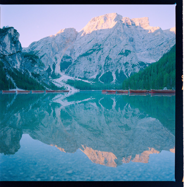 A majestic, snow-capped mountain stands silhouetted against a dusky sky, its reflection shimmering in the calm waters of a still lake, with numerous canoes resting on the shore.