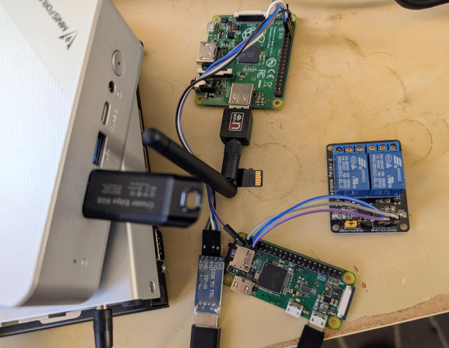 A top-down shot of a small computer setup on a tan surface with some visible coffee stains. In the upper left, a silver mini-PC is partially visible, with a black USB stick plugged into its side. Below the mini-PC, a black USB to TTL serial adapter is connected to and powering a Raspberry Pi A+. The Raspberry Pi Zero W is connected to and driving a 2-relay module. 
The Raspberry Pis are running NetBSD, the mini-PC is running llumos/SmartOS, and the two, slightly visible APU devices are running FreeBSD.