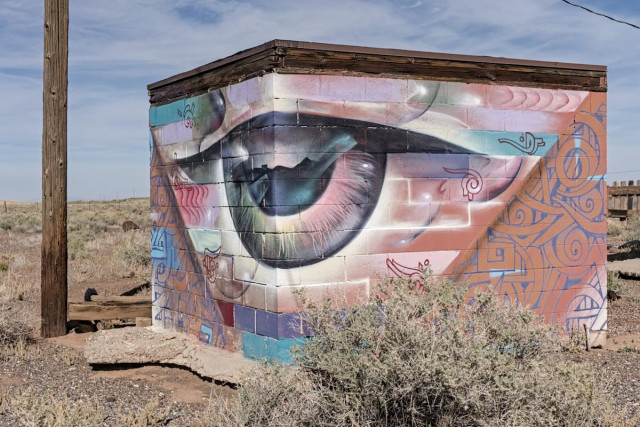Color landscape photo of street art on a small square outbuilding. The view is looking toward one corner. So wee see two sides. The corner is the center of the artwork, which is an eyeball looking out to a mountainous horizon wit a rising sun that is reflected in the dark pupil. The iris is light reds and grays. The eye socket fades into a collage of various Native symbols and geometric shapes all in light pastels colors of reds, whites, blues, and purple. A wood power pole stands to the left of the small outbuilding.