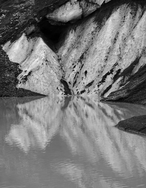 A black and white photo of a section of a glacier with a dark stripes and holes which is reflected in a calm body of water surrounded by dark rocky terrain.

