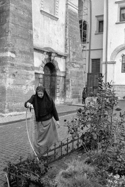 The black-and-white photograph shows a nun watering a small garden in the courtyard of a church. She wears a dark veil and long skirt, holding a hose as water gently flows over the plants. Behind her rises the stone wall of the church, with a large arched doorway and weathered masonry that speaks of age and history. In the background, a child stands quietly near the building, adding a subtle human presence to the tranquil scene. The image conveys a sense of calm devotion and simple, everyday grace within a sacred space
