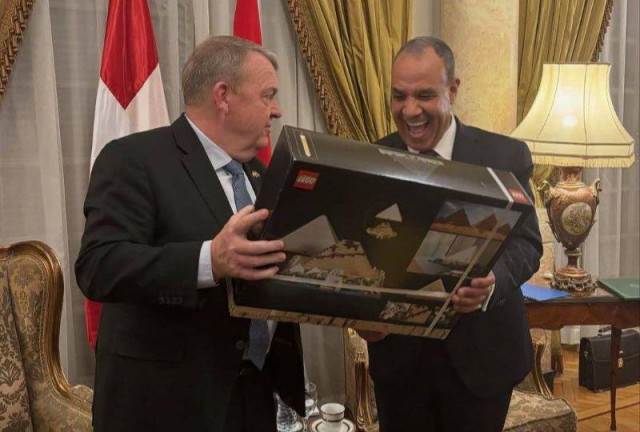 Danish minister give the Egyptian minister a Lego set of the Pyramids in a formal looking room, flags, gold, big lamp shades, the expression on the face of the Egyptian minister is just pure joy and the biggest smile ever.