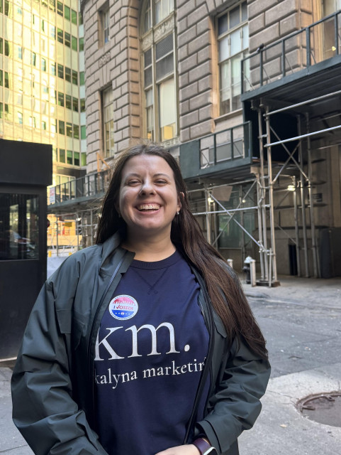 Photo of Mariya, a white woman with long brown hair, smiling in the middle of a downtown Manhattan street, showing off her “I VOTED” sticker on her shirt. 