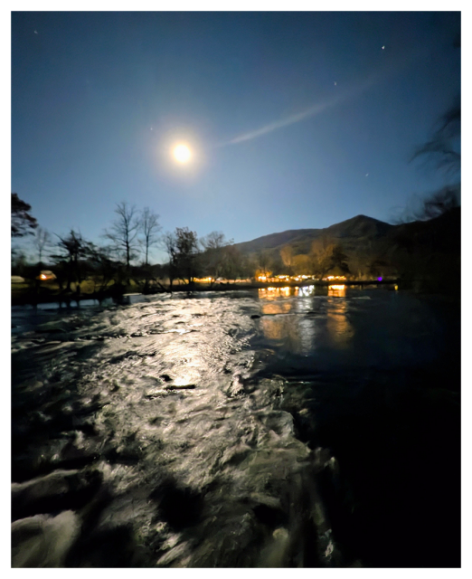 A bright full moon illuminates a gently flowing river at night. In the background, lights from buildings glow warmly among silhouetted trees, with misty mountains rising beneath a clear, starry sky. The moonlight reflects on the rippling water.