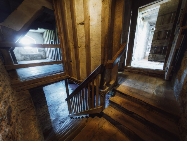 The interior of Stokesay Castle's Solar Block stairwell descends into shadow and history, with substantial timber posts and beams rendered in warm honey and amber tones creating a framework of medieval craftsmanship. Bright daylight streams through two tall windows, one on either side of the staircase, casting sharp geometric shadows across the worn wooden steps and illuminating the rendered stone walls with an ethereal blue-white glow that contrasts strikingly with the aged timber. A sturdy wooden handrail with vertical balusters guides the descent, whilst the exposed ceiling beams and structural supports overhead speak to the 14th-century construction, all contributing to the palpable sense of stepping back through centuries as one moves from the comparatively brighter ground floor towards the dim cellar below.
