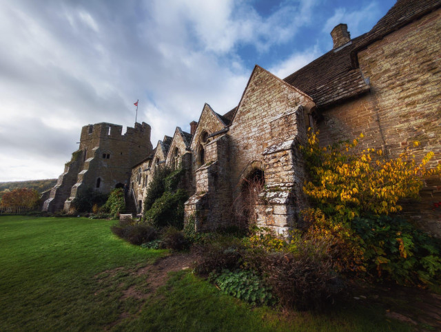 Stokesay Castle presents a striking façade of honey-coloured sandstone beneath a dramatic autumn sky, its crenellated towers and steeply pitched slate roofs speaking to centuries of border fortification in the Welsh Marches. The castle's south-facing elevation reveals the distinctive Early English Gothic architecture characteristic of the 13th century, with lancet windows piercing the substantial walls and a commanding square tower flying the Union flag rising prominently from the left. Golden-hued ivy and autumn foliage in shades of amber and bronze cling to the weathered stone in the foreground, softening the fortress's formidable appearance, whilst the lush green sward stretches before the building beneath a brooding sky scattered with white clouds, creating a quintessentially English pastoral backdrop that belies the castle's martial heritage as one of the finest surviving fortified manor houses in England.
