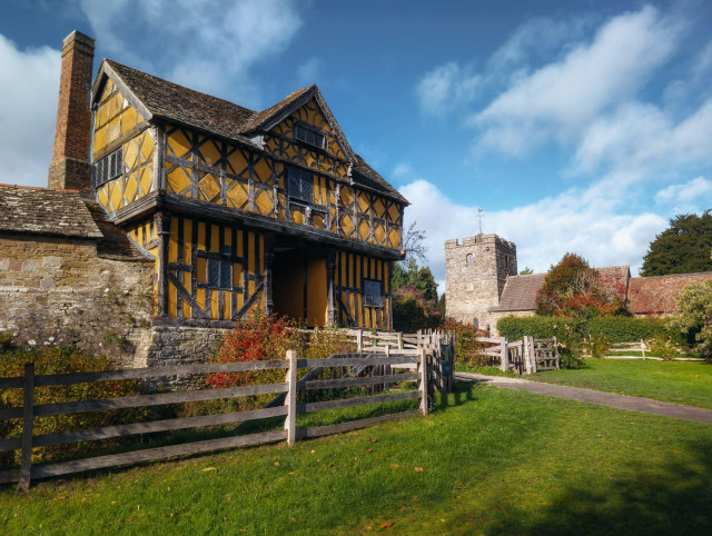 The timber-framed gatehouse of Stokesay Castle commands attention in vivid ochre and black half-timbering, its steeply pitched gables and prominent brick chimney silhouetted against a brilliant blue sky dotted with white clouds, whilst weathered wooden fencing in the foreground suggests the domestic pastoral character of this 16th-century structure. The jettied construction of the upper storeys displays the distinctive diamond and geometric patterning characteristic of Elizabethan timber-framing, with rows of vertical and diagonal bracing creating bold visual rhythms across the ochre-rendered infill panels. Beyond the gatehouse, the stone Church of Saint John the Baptist rises with its sturdy tower and russet-tiled roofing, anchoring the composition in the middle distance amongst autumn-tinged vegetation, whilst the meticulously maintained green sward and the substantial stone foundations visible to the left ground the scene in the bucolic countryside of the Welsh Marches, creating a harmonious ensemble of medieval religious and domestic architecture set within the timeless English landscape.