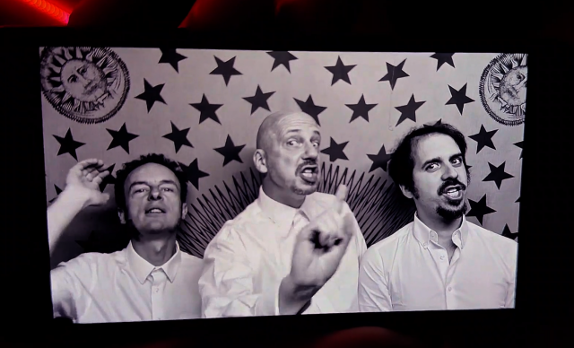 A picture of a phone screen showing a black and white image of three white old guys in white shirts, mid-sentence, gesticulating