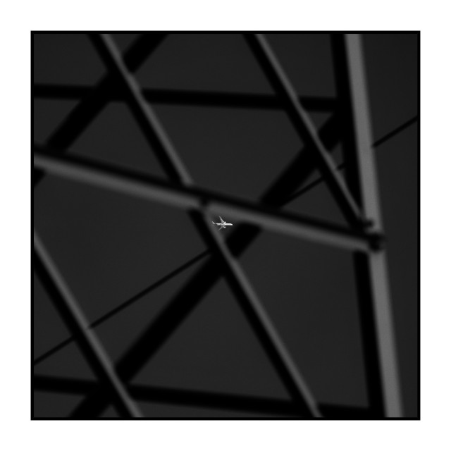 Square black-and-white photograph. The image shows the structure of a metal tower, with diagonal and vertical beams crossing the frame. The beams are out of focus and form intersecting lines against a dark sky. In the center of the image, sharply focused, a small airplane is visible flying through one of the open spaces between the beams.