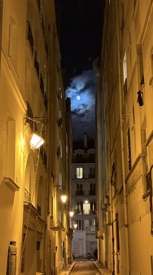 Photo d'une rue étroite à Paris, immeubles de chaque côté lumière jaune tamisée, immeuble au milieu au fond de la rue et vue sur la pleine lune entourée de nuages dans le ciel