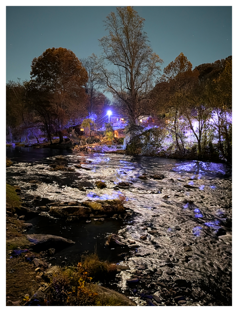 A gently flowing, narrow river runs through a wooded area at night. Trees with autumn foliage line the banks, and blue lights from houses in the background reflect on the water. the sky is clear.
