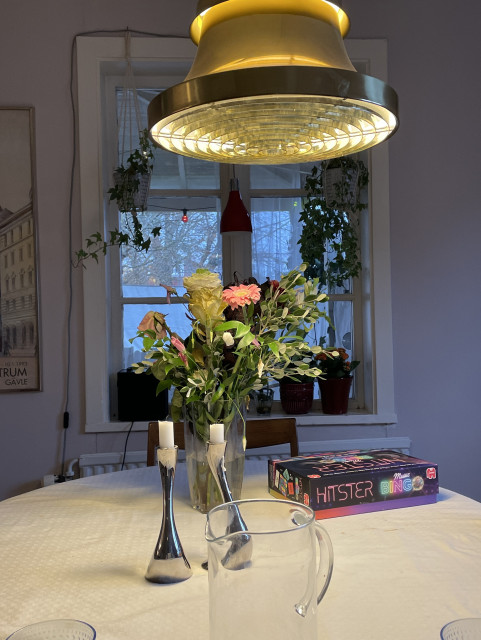 A dining table topped with a tablecloth features a vase of mixed flowers, two candle holders, and a game box labeled "Hitster Music Bingo." A sleek, modern pendant light hangs above the table, with a backdrop of windows displaying plants.