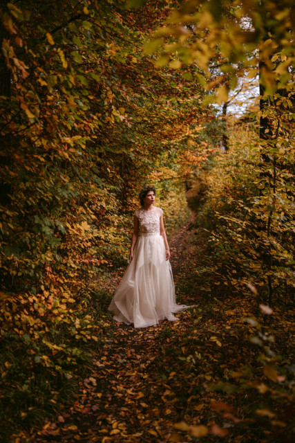 A bride in an elegant white beaded bodice gown with a flowing wedding dress stands centered in a narrow autumn forest tunnel, shot by European Elopement & Wedding Photographer Sturmsucht. Dense hedges and shrubs line both sides, their foliage in shades of golden yellow, orange, and rust. The ground is carpeted with fallen leaves. Dappled sunlight filters through the canopy above, creating a warm, golden glow. The woman faces slightly left, her dark hair visible against the pale dress. The intimate pathway creates a peaceful, contemplative autumn atmosphere.