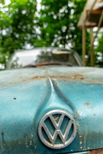Auf dem Bild ist die Vorderseite eines alten, rostigen VW 1500 zu sehen, erkennbar an der Form der Haube und dem markanten VW-Logo. Die Lackierung des Autos ist türkis und weist deutliche Rostspuren auf. Im unscharfen Hintergrund sind Bäume und eine Holzstruktur zu erkennen.