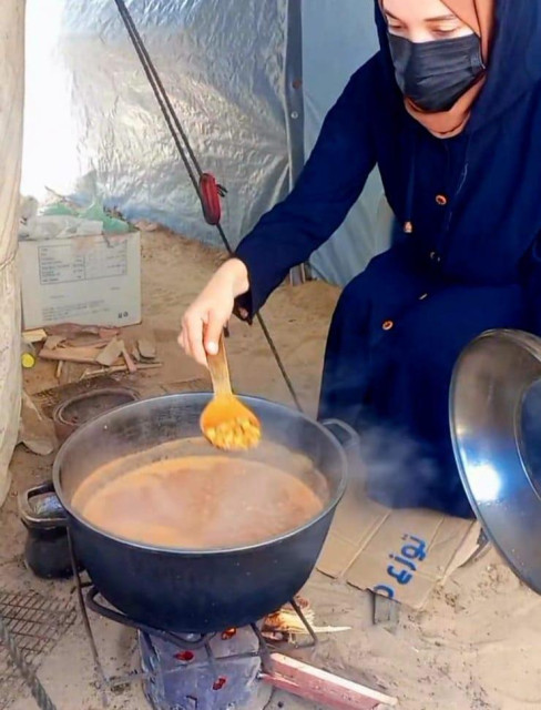 a woman wearing a mask cooks with a pot on an open fire, dishing out what look like beans with a wooden spoon from a stew 💔