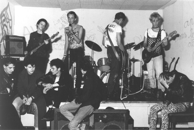 Schwarzweiss Foto eines Bühnenauftritts einer sehr jungen Punkband. Vier junge Männer stehen auf einer Bühne und halten Gitarren oder Mikrofone - teilweise wirken sie sehr mit sich selbst beschäftigt. Im Vordergrund sitzen mehrere Personen in punkiger Aufmachung. Sie machen einen gelangweilten Eindruck