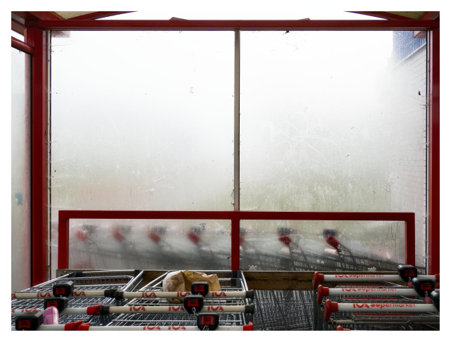 En liggande bild på ett fönster på en plats för vagnar vid en ICA-affär. Det är disigt och vagnarna på andra sidan fönstret är oskarpa pga diset och fönstret. Röda balkar ramar in vagnarna, en papperspåse och en rosa burk ligger på ett par av vagnarna.