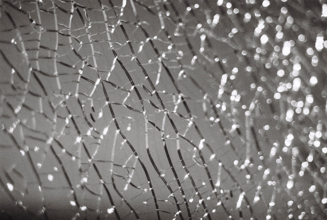 Close-up black-and-white photograph of a shattered glass surface. A web of cracks covers the entire surface like glimmering silver threads. The reflections in the background almost look like the sun shining on waves in the sea. Pretty, but broken.