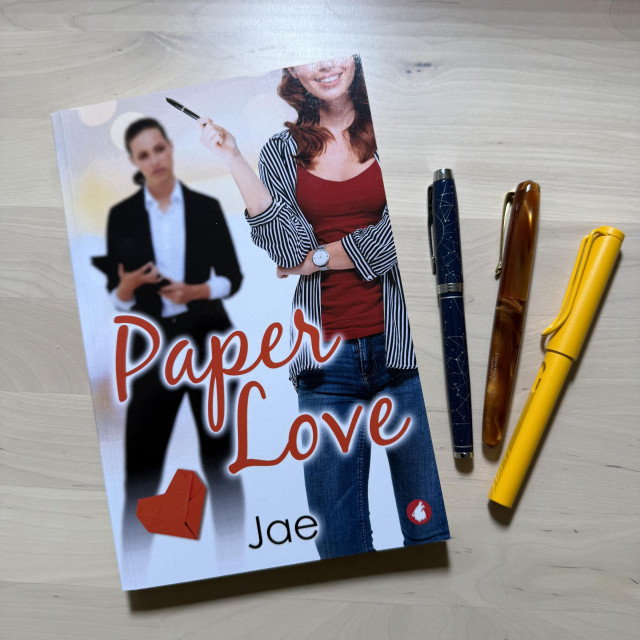 Shown is a paperback copy of Paper Love by Jae, two women, one holding an iPad, the other holding a fountain pen. Next to the book are three fountain pens (blue, copper, and yellow). 