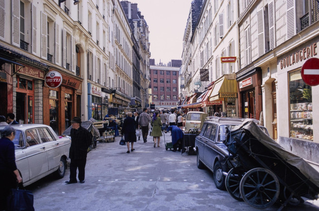 Des passants se promènent dans une rue commerçante, une charrette à bras est garée au premier plan