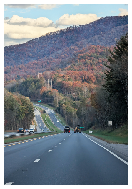 A scenic, four-lane highway curves through rolling hills covered in autumn-colored trees. Several vehicles drive on the road, in both directions and a speed limit sign reads “60.” In the background, a large forested mountain rises under a partly cloudy sky