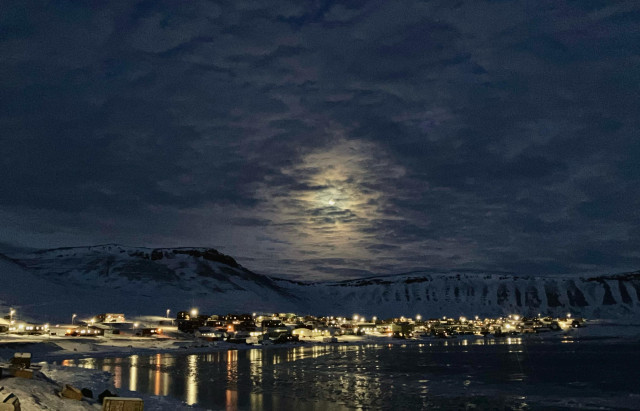 A mostly full moon scatters its light among the clouds. Below it a ring of hills hold a small lit town in their arms. The lights of town reflected in the water and ice of the bay. 