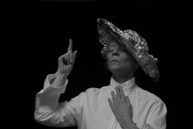 Ett porträtt av en kvinna i en foiliehatt och med en högt knäppt vit sjkorta med ena handen upp mot kragen och hållandes den andra upp i luften med ett förmanande finger. Hon håller huvudet högt, blundar och har ett bistert ansiktsuttryck. Fotot är svartvitt och hon sticker tydligt ut mot den kolsvarta bakgrunden.  