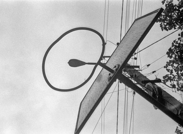 The black and white photograph shows a basketball hoop and backboard viewed from below. The hoop’s circular outline frames a streetlight in the background, creating an unexpected visual overlap. Power lines stretch diagonally across the sky, adding a web of geometric lines around the composition. The image has a gritty, film-grain texture that enhances its raw, urban atmosphere and sense of quiet abstraction.
