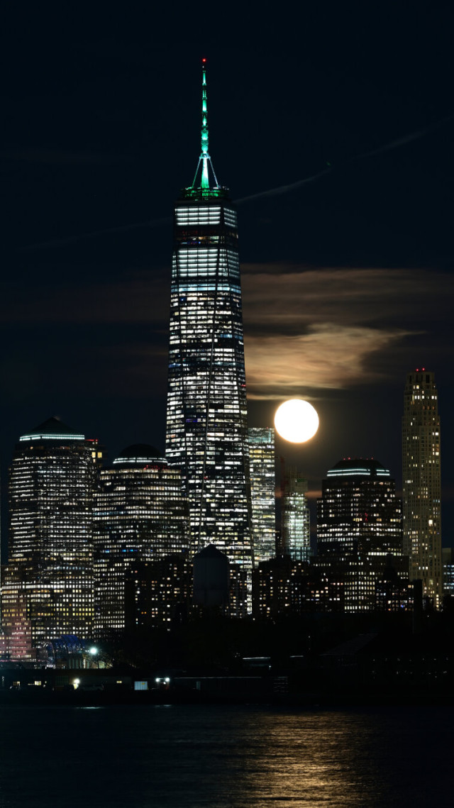 A full moon rises from behind the buildings of lower Manhattan, most notably One World Trade Center, as seen from New Jersey.