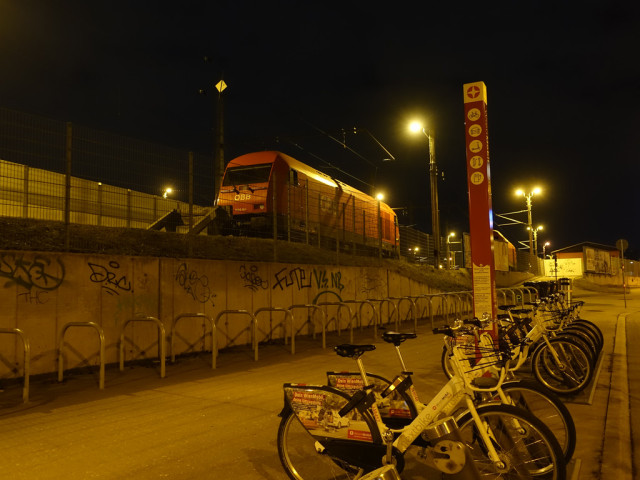 Gehsteig mit Leihradstation bei Nacht, links davon in erhöhter Position eine 2016-Lokomotive der ÖBB auf einem Abstellgleis.