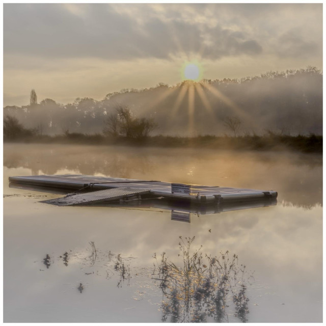 A serene landscape featuring a calm lake at sunrise. A floating dock is visible in the foreground, surrounded by mist and soft reflections in the water. Sun rays break through the clouds, illuminating the tranquil scene. Trees are faintly seen in the background.