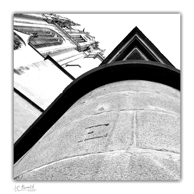 Square black-and-white image of Frankfurt Cathedral. The view goes up a round corner of the building made of rough stones with an angular finish above it and an engraved ‘E’. To the left, you can see the façade of the Gothic cathedral with its tall windows reaching up into the white sky.