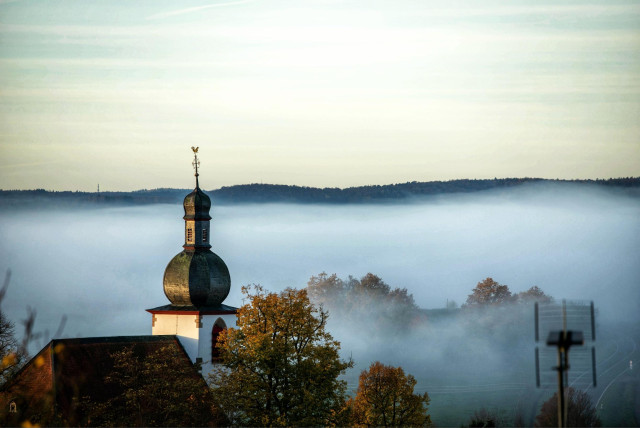 Auf dem Bild ragt eine Kirche mit ihrem markanten Zwiebelturm aus einer Nebeldecke, die sich über die umliegende Landschaft gelegt hat. Im Vordergrund sind Bäume zu erkennen, und im Hintergrund sind bewaldete Hügel sichtbar.