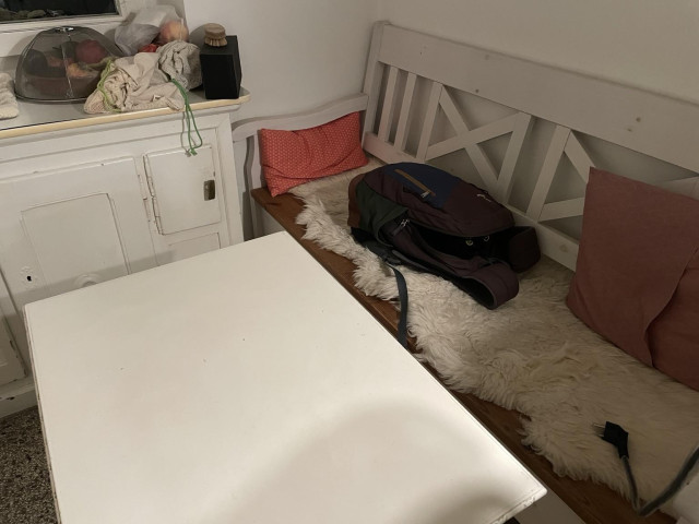 A cozy indoor space featuring a white wooden table and bench with pink cushions. A backpack is resting on the bench, partially open, with an extension cord nearby. A cupboard holds a covered dish of fruits, and a few cloth bags are tucked beside it. No cat to be spotted anywhere…except…these eyes?!👀 