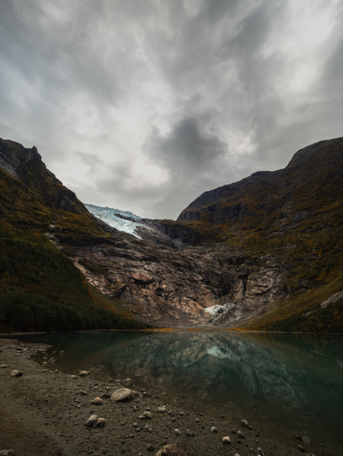 A dramatic Norwegian mountain valley with a calm turquoise glacial lake reflecting steep, moss-covered slopes on both sides. Exposed dark rock faces and grey stone create a narrow U-shaped valley. A pale blue-white glacier is visible nestled high between the peaks in the background. The foreground shows a pebbly beach with scattered stones. An overcast sky with thick grey and white clouds creates a moody atmosphere over this remote, pristine wilderness landscape.