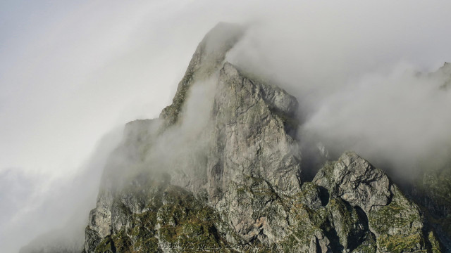 A colour photo of a mountain top surrounded by dense cloud. The peak has two distinct tops and there is cloud between them, giving the impression of being caught by them in a pinch. The rock is mostly grey but some green vegetation can be seen on horizontal ledges.