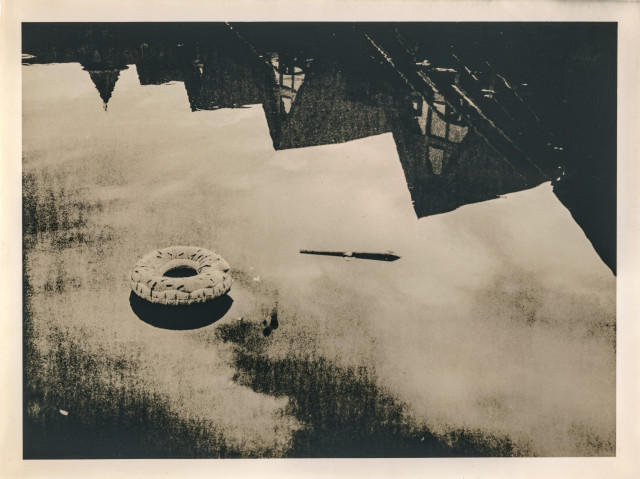 This lith print depicts a quiet, reflective water surface with a floating rubber ring and a wooden stick. The upper part of the image shows the dark, angular reflections of buildings on the water, their sharp forms contrasting with the soft texture of the surface. The print’s gritty texture, muted tones, and high contrast give it a dreamlike, almost melancholic atmosphere — a sense of stillness and abandonment. The balance between the floating objects and the distorted reflections creates a poetic tension between reality and illusion.
