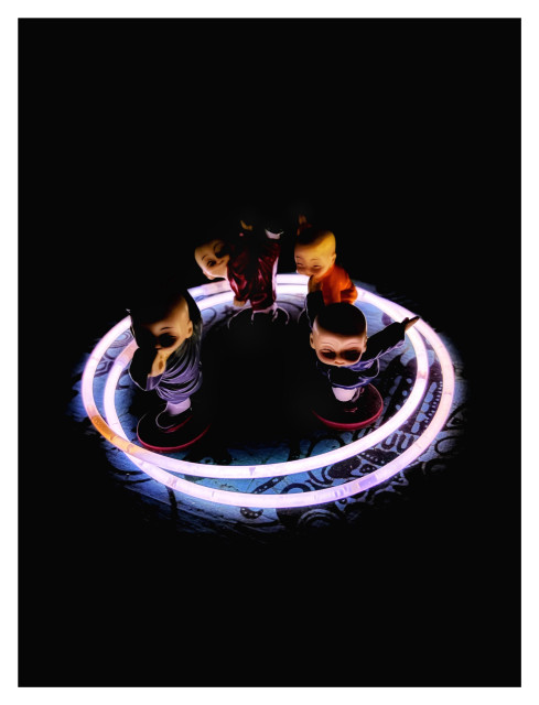 Photo of four miniature ceramic figurines of bald, baby-faced monks in different colored tunics and striking different martial arts poses. They’re grouped together inside two overlapping circles created by concert glow sticks configured into rings, on a tabletop covered in blue batik fabric. The only light comes from the glowing rings, which create striking contrasts of light and shadow on the figurines while leaving the rest of the image in darkness.