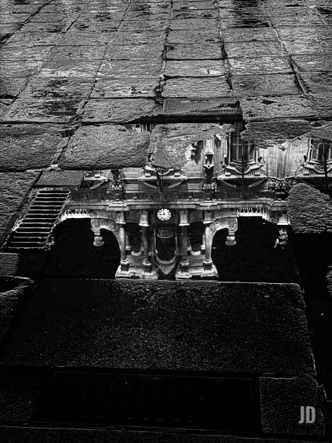 Fotografía en blanco y negro que captura el reflejo de un edificio ornamentado (que parece tener una fachada barroca o similar, con columnas, arcos y un reloj en el centro) sobre una superficie mojada y adoquinada.
El reflejo es el elemento principal y más claro de la composición, apareciendo en un charco entre las piedras oscuras del pavimento. La perspectiva es peculiar, haciendo que el reflejo parezca casi un agujero o portal en el suelo que muestra la estructura del edificio invertida.
En resumen, es una imagen de contraste y simetría que juega con el reflejo de una arquitectura histórica sobre un suelo mojado.