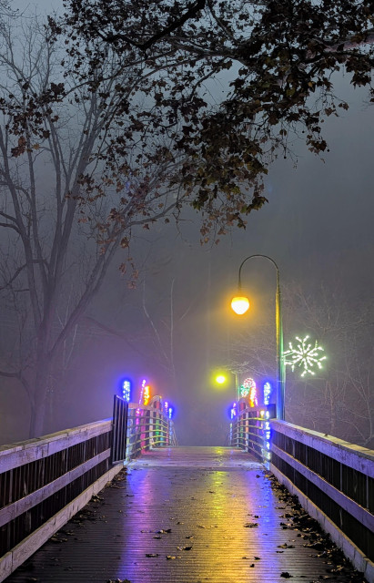 foggy morning before sunrise. The view across a wooden bridge with lamp post light at the end and holiday decorations in neon colors on both sides. The golden leaves of a tree hang over them. The background is obscured by fog.